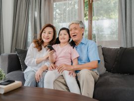 Asian grandparents watch TV with granddaughter at home. Senior Chinese, grandfather and grandmother happy using family time relax with young girl kid lying on sofa in living room concept.