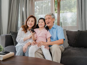 Asian grandparents watch TV with granddaughter at home. Senior Chinese, grandfather and grandmother happy using family time relax with young girl kid lying on sofa in living room concept.