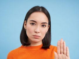 Close up portrait of serious asian woman, shows stops sign, disapproves something, refuses or rejects, stands over blue background.
