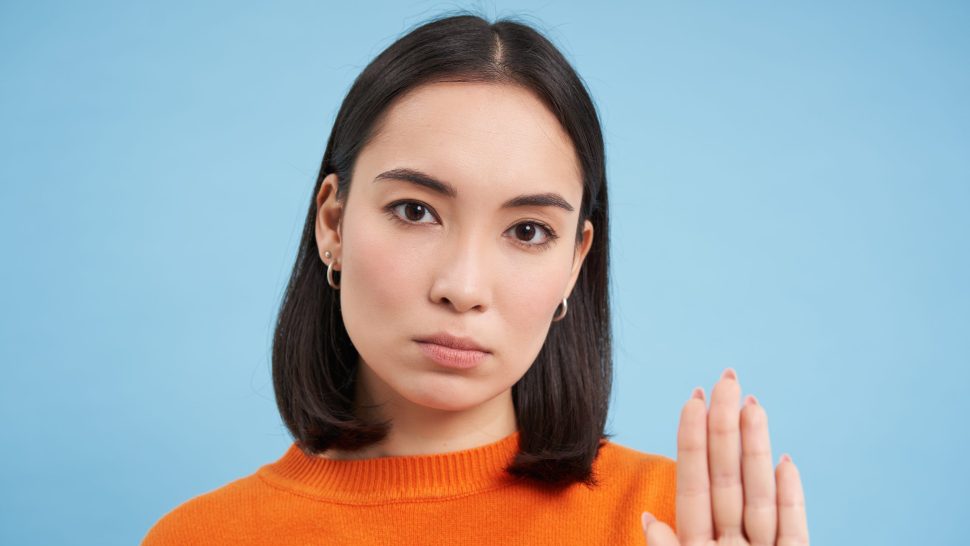 Close up portrait of serious asian woman, shows stops sign, disapproves something, refuses or rejects, stands over blue background.