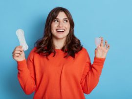 Happy excited dark haired female looking away with with toothy smile, comparing menstrual cap and cotton pad, copy space for promotion, isolated over blue background.