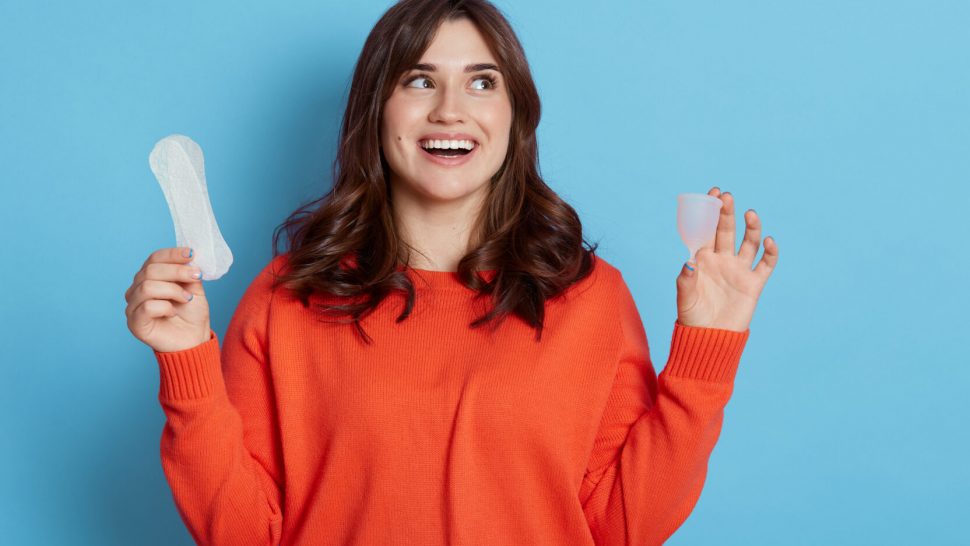 Happy excited dark haired female looking away with with toothy smile, comparing menstrual cap and cotton pad, copy space for promotion, isolated over blue background.