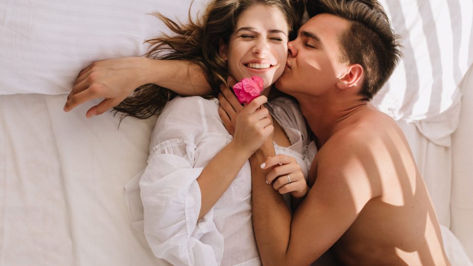 Overhead portrait of tanned sleepy boy with trendy hairstyle kissing his cute girlfriend, lying in bed with eyes closed. Adorable young woman in blouse holding pink flower and enjoying morning in bed