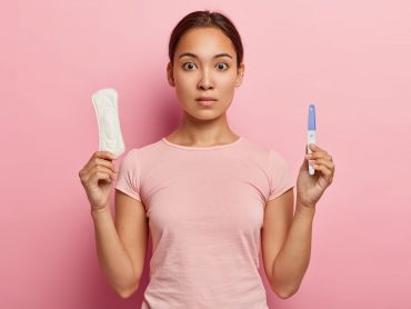 Photo of attractive Asian woman holds cotton tampon and sanitary napkin, has surprised look, monthly menstrual cycle, wears pink t shirt. Protective care for womens health. Fertilization concept