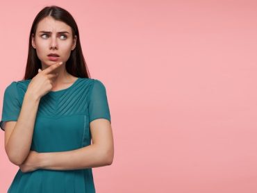 portrait-attractive-thinking-girl-with-long-brunette-hair-folding-hands-chest-touching-her-chin-watching-right-copy-space-pastel-pink-wall-wearing-emerald-dress_295783-9227