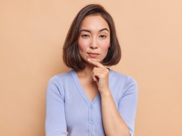 portrait-serious-dark-haired-asian-woman-keeps-finger-chin-looks-mysteriously-front-considers-something-dressed-blue-jumper-isolated-brown-wall_273609-53356