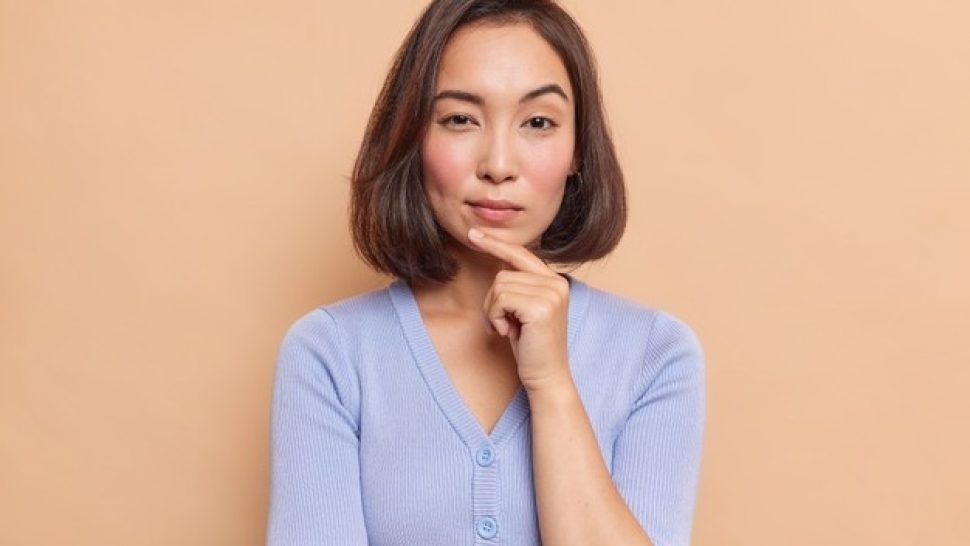 portrait-serious-dark-haired-asian-woman-keeps-finger-chin-looks-mysteriously-front-considers-something-dressed-blue-jumper-isolated-brown-wall_273609-53356