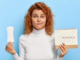 Puzzled ginger woman holds sanitary napkin and menstruation calendar with marked red days, suffers from period cramps, controls her women health, has periods this week, poses over blue background
