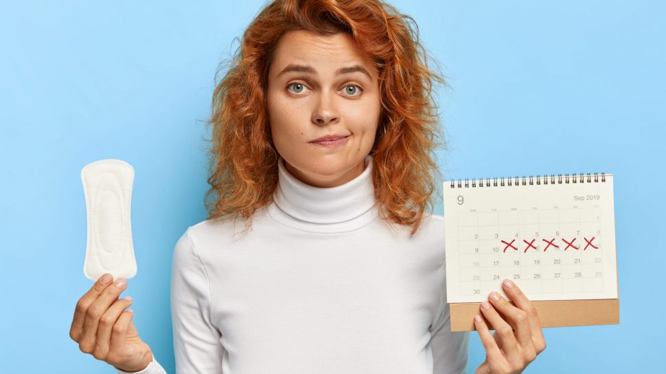 Puzzled ginger woman holds sanitary napkin and menstruation calendar with marked red days, suffers from period cramps, controls her women health, has periods this week, poses over blue background