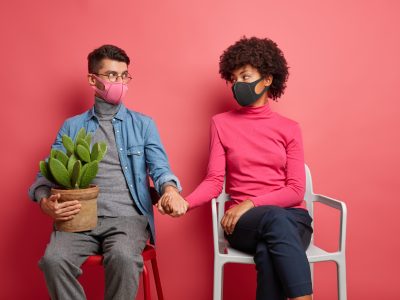 Serious husband and wife infected with coronavirus support each other hold hands wear protective face masks sit on chairs wear casual clothing spend time at home isolated over pink background