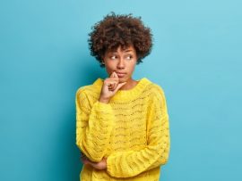 serious-pensive-woman-with-afro-hair-looks-away-stands-thoughtful-pose-thinks-about-problems-difficulties-dressed-warm-yellow-sweater-isolated-blue-wall-makes-decision_273609-44649