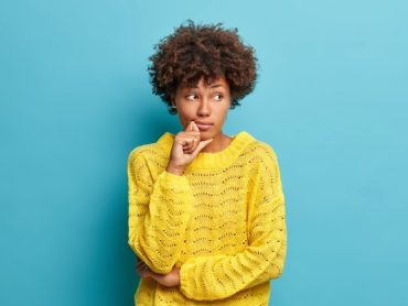 serious-pensive-woman-with-afro-hair-looks-away-stands-thoughtful-pose-thinks-about-problems-difficulties-dressed-warm-yellow-sweater-isolated-blue-wall-makes-decision_273609-44649