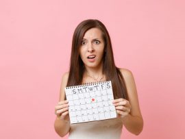 Shocked concerned bride woman in white wedding dress holding female periods calendar for checking menstruation days isolated on pink background. Medical, healthcare, gynecological concept. Copy space