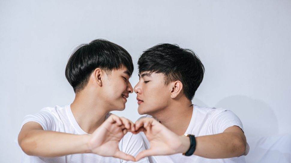 Two loving young men look at each other and make a hand of love symbol.