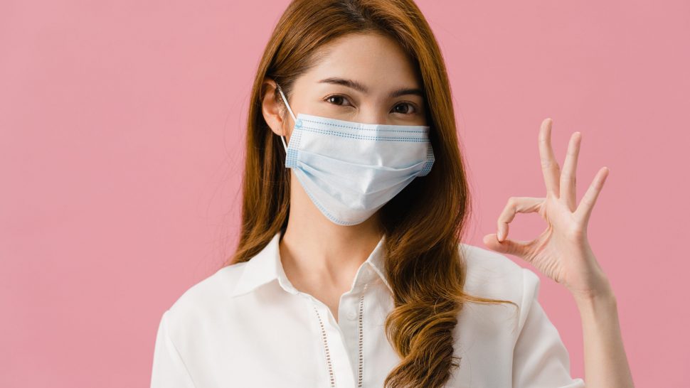 Young Asia girl wearing medical face mask gesturing ok sign with dressed in casual cloth and look at camera isolated on pink background. Self-isolation, social distancing, quarantine for corona virus.