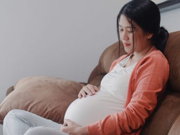 Young Asian Pregnant woman holding her belly talking with her child. Mom feeling happy smiling positive and peaceful while take care baby, pregnancy lying on sofa in living room at home concept.