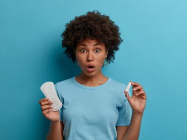 Photo of curly haired stunned emotive woman holds sanitary napkin and tampon, has menstrual cycle. Emotional dark skinned female model poses with feminine hygiene products. Women health concept