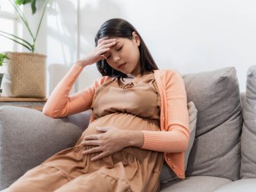 Young pregnant asian woman has suffering from headache sitting on sofa. Pregnancy symptoms